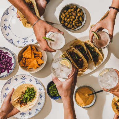 Table with mexican food on it and people cheersing their glasses for dinner.